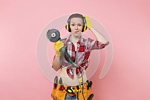 Confused energy handyman woman in yellow gloves, noise insulated headphones, kit tools belt full of instruments holding