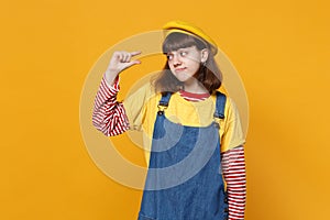 Confused girl teenager in french beret, denim sundress gesturing demonstrating size with workspace isolated on yellow