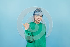 Confused hipster girl with blue hair stands on a blue background and shows a finger to the side with a sad face