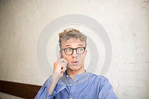 Confused hipster man talking via mobile phone, sitting in modern interior against brick wall.