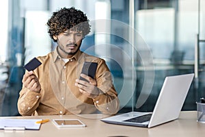 Confused hispanic man looking clueless at cellular phone while holding bank card and sitting at cabinet with laptop