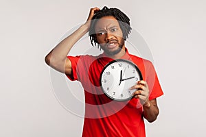 Confused man with dreadlocks wearing red T-shirt, rubbing his head holding in hands big wallclock, has no time , worried about photo