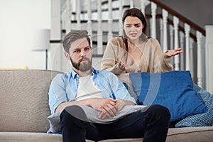 Confused man sits on the couch while his girlfriend yells and quarrels with him at home. Big family problems.