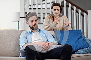 Confused man sits on the couch while his girlfriend yells and quarrels with him at home. Big family problems.