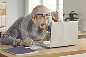 Confused mature man who has poor eyesight and works on laptop at home wearing glasses.
