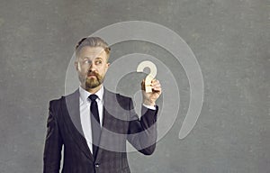 Confused puzzled businessman holding wooden question mark standing on gray on background.