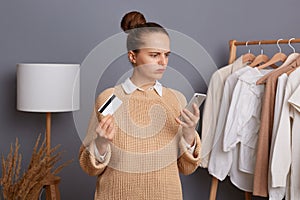 Confused puzzled woman in beige sweater stands against gray wall with clothes hang in wardrobe on rack, holding cell phone and