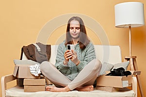 Confused puzzled woman opening boxes of postal parcel with smartphone sitting on sofa against beige wall having troubles with