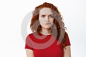 Confused redhead girl frowning and look with doubtful face, feeling unsure or puzzled, standing pensive in red t-shirt