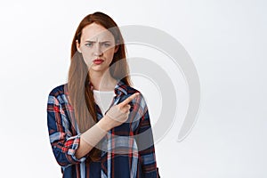 Confused redhead girl frowns suspicious, pointing at upper right corner with displeased face expression, showing