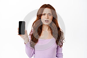 Confused redhead woman shows something bad on screen of mobile phone, frowning and staring puzzled or frustated