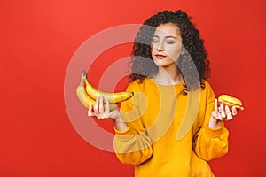 Confused sad upset curly woman can`t make a choice between healthy and unhealthy food, she is holding fruits and donut on her