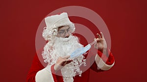 Confused, Santa is holding a protective mask and does not know how to dress against the background of a red wall. Puzzled Santa