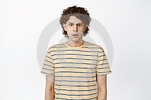 Confused and shocked curly guy raising eyebrow, looking startled and standing in stupor, standing over white background