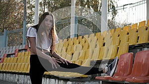 Confused slim young female in sportswear stretching legs at tribune of stadium