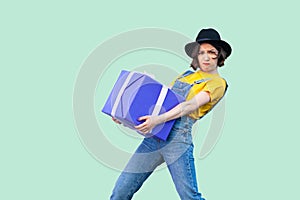 Confused strong young girl in fashionable hipster wear in denim overalls and black hat standing and trying to hold giant big heavy