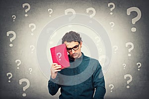 Confused student guy looks down upset, holding a book with question mark and different interrogation symbols around head. Concept