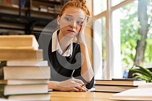 Confused tired redhead lady student sitting at the table with books in library writing notes