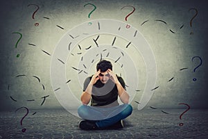 Confused and tired young man seated on the floor keeps hands to forehead looking down thoughtful surrounded by different tasks to