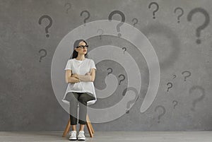 Confused woman sitting on chair and looking at lots of question marks on grey background
