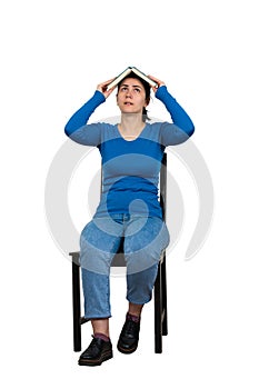 Confused woman student, seated on a chair, keeps open book over head isolated on white. Difficult task concept, discontent girl