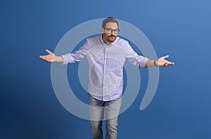 Confused young businessman in glasses shrugging shoulders while standing on isolated blue background
