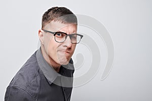 Confused young man with glasses squinting and frowning against a gray background