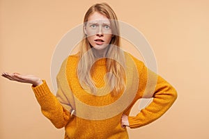 Confused young pretty redhead female frowning her eyebrows and raising perplexedly palm while standing over beige background in