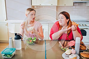 Confused young slim model eat farmer cheese at table. She look at plus size model who eats burgers and junk food. Happy