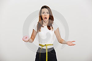 Confused young woman with centimeter around waist and donut in hand.