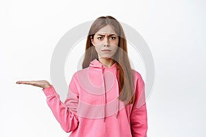 Confused young woman frowning with judgement, holding something on hand, display product on her palm against copy space