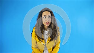 Confused young woman in yellow jacket in studio with blue background.