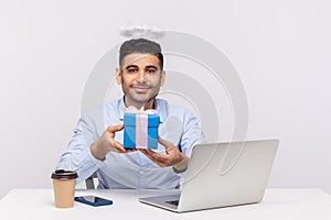 Congratulations! Happy angelic businessman with nimbus on head holding gift box, sitting at laptop workplace
