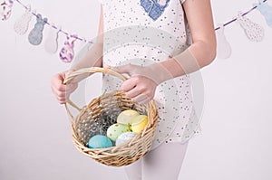 Congratulations on happy Easter: the girl is holding a basket with painted eggs by the white wall.