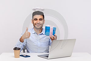 Congratulations! Joyful angelic businessman with nimbus on head showing gift box and thumbs up, sitting at laptop