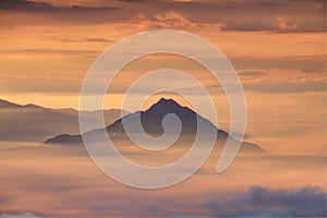 Conical peak emerges from sea of clouds and orange mist at dawn