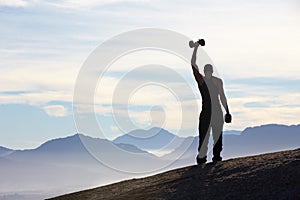 Conquering his world. Rearview of a man standing on a mountain top holding a dumbell high above his head.