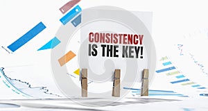 CONSISTENCY IS KEY text on paper sheet with chart, dice, spectacles, pen, laptop and blue and yellow push pin on wooden table -