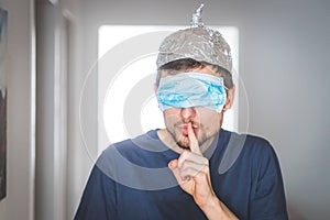Conspiracy theory concept: Young man with face mask over the eyes and aluminum hat is making a psst! gesture