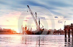 Construction of an automobile bridge with cranes and pouring concrete over the river in the rays of the setting sun