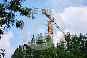 A construction crane works against a background of blue sky and green trees. The construction of a high-rise building with an