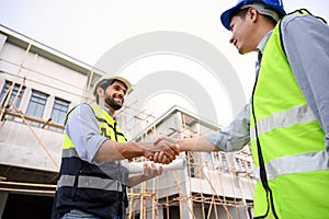 Construction engineer and architect with blueprint shaking hands while standing on construction site. Home building concept