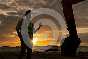 Construction engineer holding radio communications with backhoe in construction site by the sea at sunset.