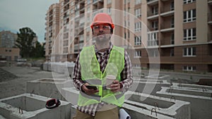 Construction industry worker texting during lunch break. Helmeted handyman chatting and using smartphone during coffee