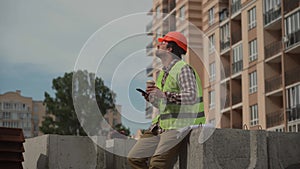 Construction industry worker texting during lunch break. Helmeted handyman chatting and using smartphone during coffee
