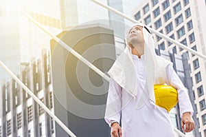 Construction and maintenance concept, Arab Middle Eastern architect holding yellow helmet and checking tablet PC with city backgro