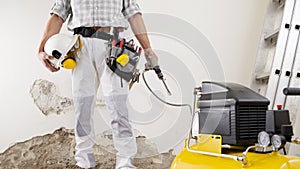 Construction man worker wearing a tool bag belt, helmet with headphones and pneumatic air hammer chisel in his hands, compressor