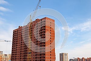 Construction of a multi-storey brick residential building of red color next to which stands a crane for lifting goods and