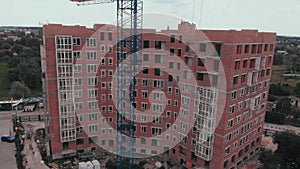 Construction of a multi-storey brick residential building of red color next to which stands a crane for lifting goods