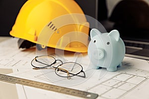 Construction safety helmet and piggy bank with blueprint, glasses and laptop on table. Building theme